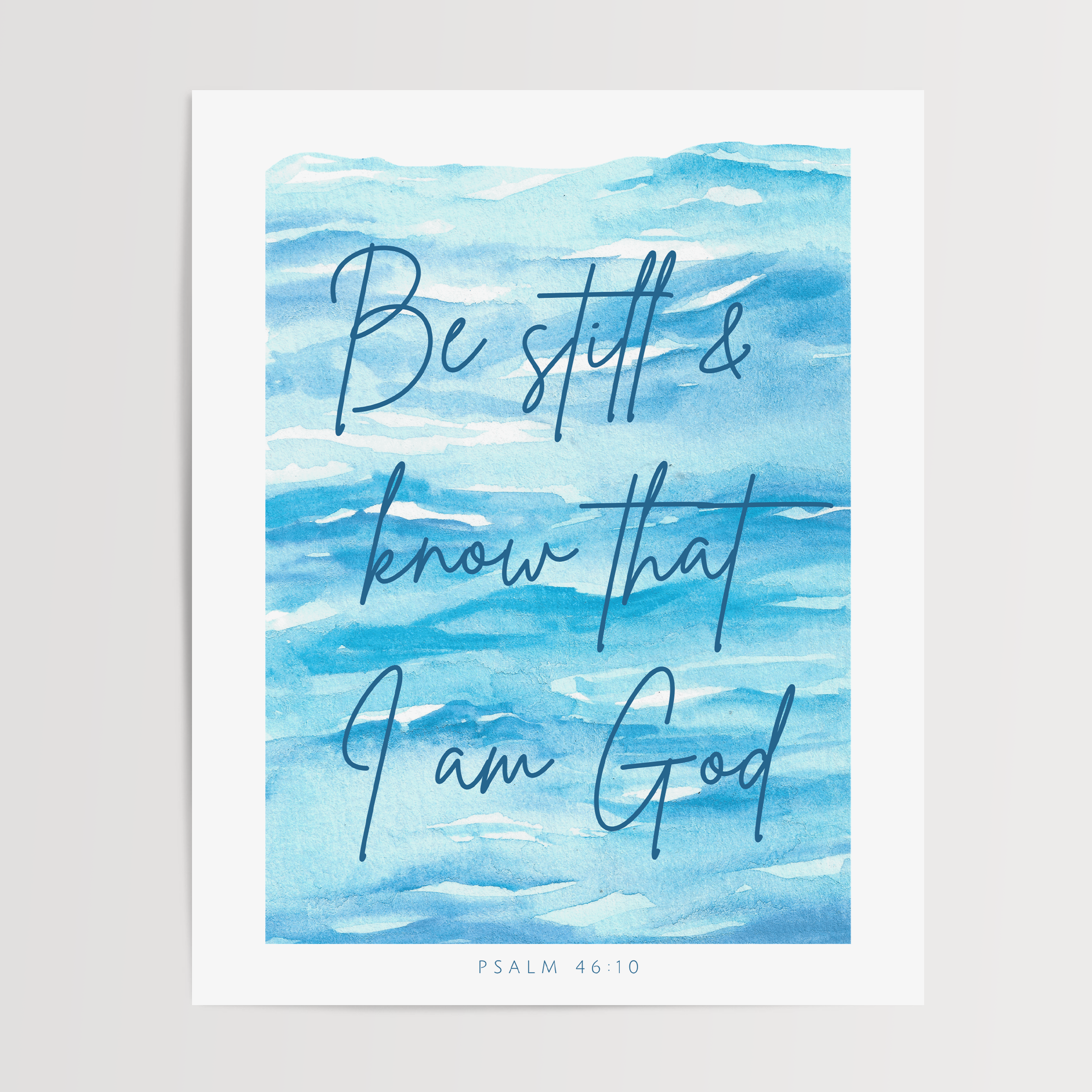A vertical art print featuring a watercolor blue ocean scene with the hand-lettered quote 'Be still & know that I am God' and 'PSALM 46:10' at the bottom, shown against a light neutral background.