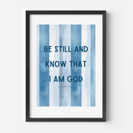 Framed wall art featuring the text 'BE STILL AND KNOW THAT I AM GOD' from Psalm 46:4 in dark blue lettering over vertical watercolor-style blue and white stripes, shown in a black frame against a light gray wall.