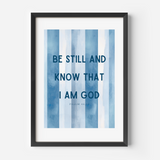 Framed wall art featuring the text 'BE STILL AND KNOW THAT I AM GOD' from Psalm 46:4 in dark blue lettering over vertical watercolor-style blue and white stripes, shown in a black frame against a light gray wall.