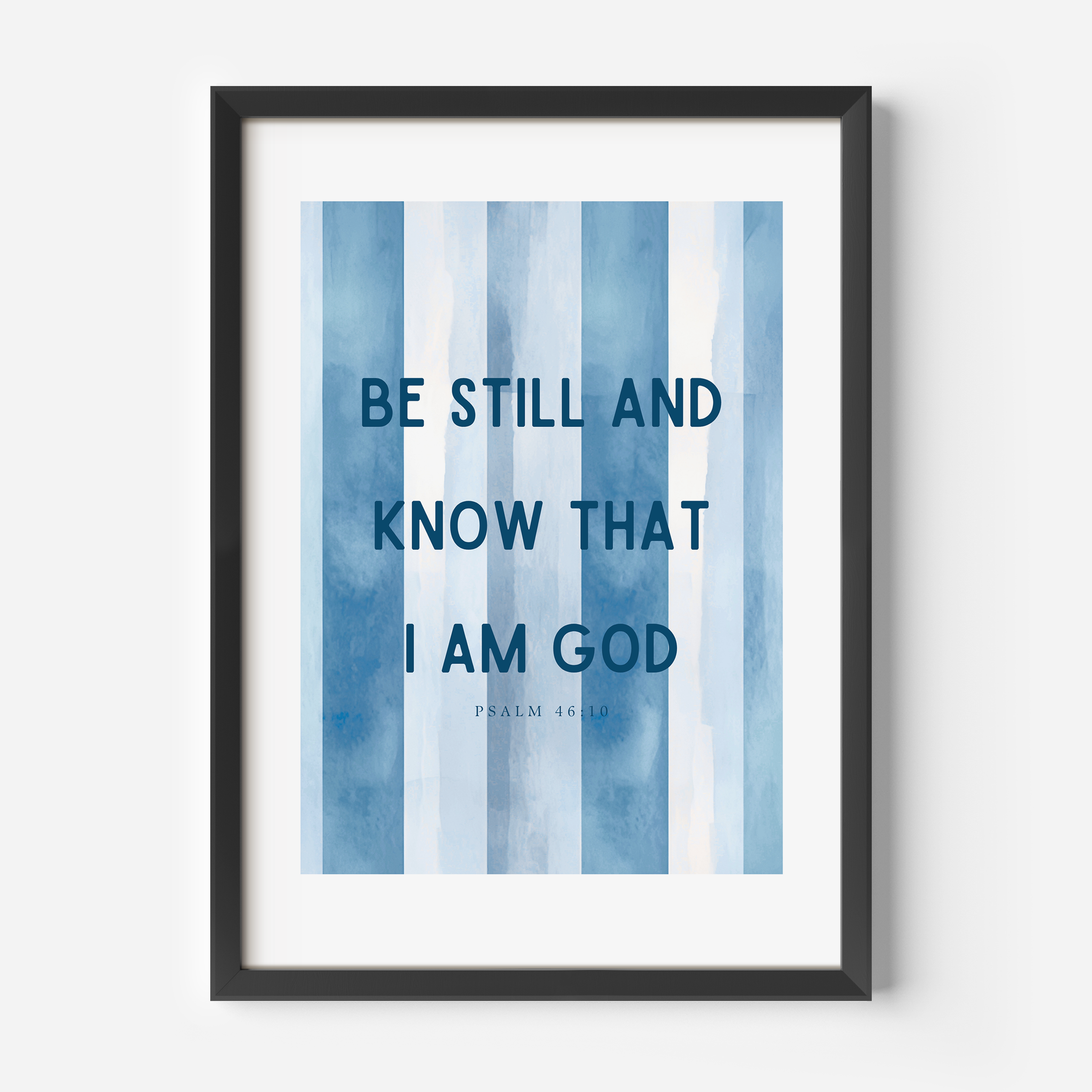 Framed wall art featuring the text 'BE STILL AND KNOW THAT I AM GOD' from Psalm 46:4 in dark blue lettering over vertical watercolor-style blue and white stripes, shown in a black frame against a light gray wall.