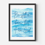 A framed art print featuring a blue watercolor background with the hand-lettered text 'Be still & know that I am God' and 'PSALM 46:10' at the bottom, shown in a black frame against a light gray wall.