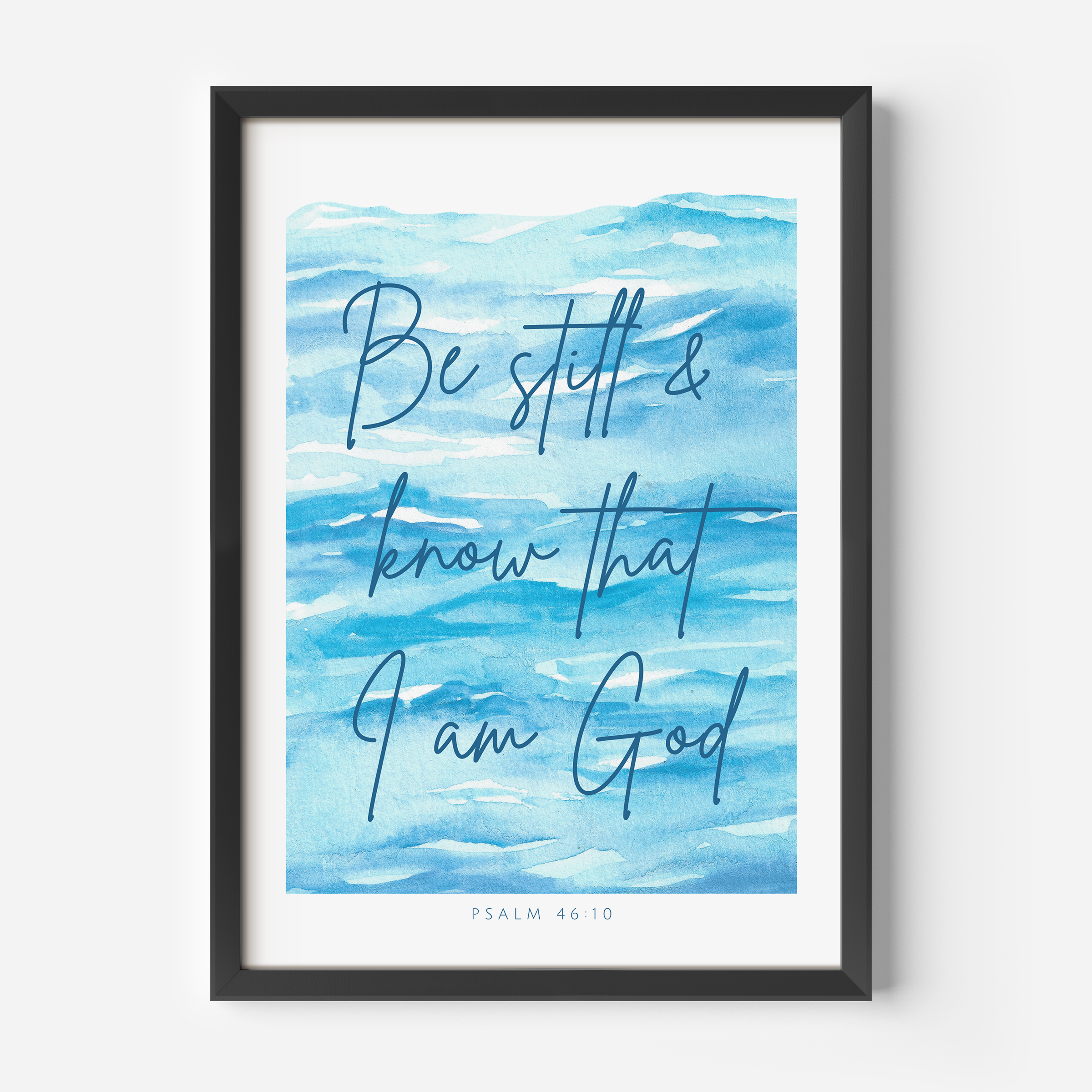 A framed art print featuring a blue watercolor background with the hand-lettered text 'Be still & know that I am God' and 'PSALM 46:10' at the bottom, shown in a black frame against a light gray wall.