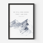 Framed wall art featuring the text 'BE STILL AND KNOW THAT I AM GOD' and 'PSALM 46:10' above a soft gray watercolor mountain illustration on a white background, shown in a black frame against a light gray wall.