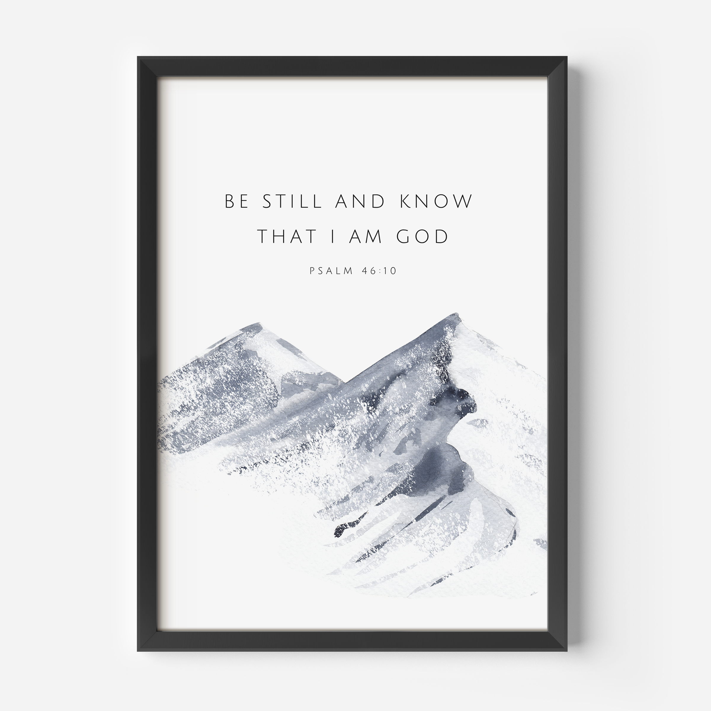 Framed wall art featuring the text 'BE STILL AND KNOW THAT I AM GOD' and 'PSALM 46:10' above a soft gray watercolor mountain illustration on a white background, shown in a black frame against a light gray wall.