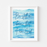 Framed wall art featuring the Bible verse 'Be still & know that I am God' in dark blue script over a watercolor-style blue background, shown in a white frame against a light gray wall.