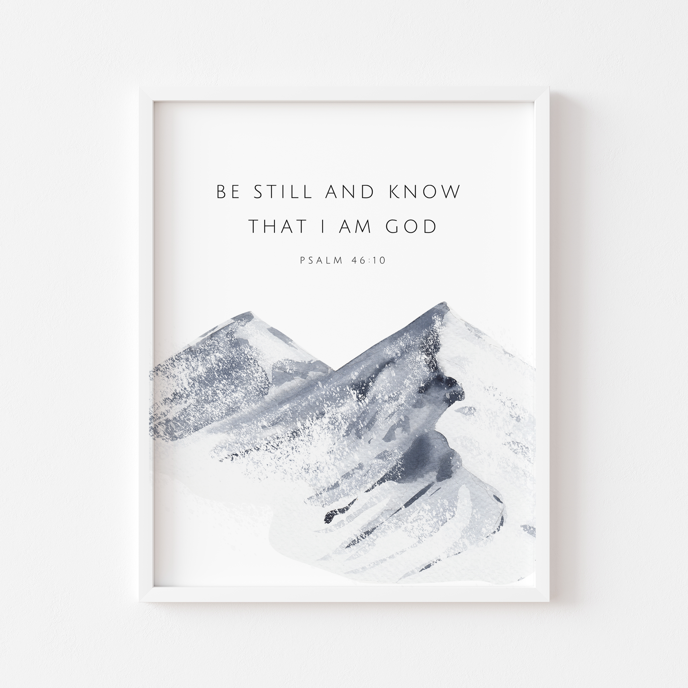 A framed wall art print featuring the text 'BE STILL AND KNOW THAT I AM GOD' and 'PSALM 46:10' above an abstract watercolor mountain scene in white, gray, and blue-gray tones, shown against a light neutral wall.