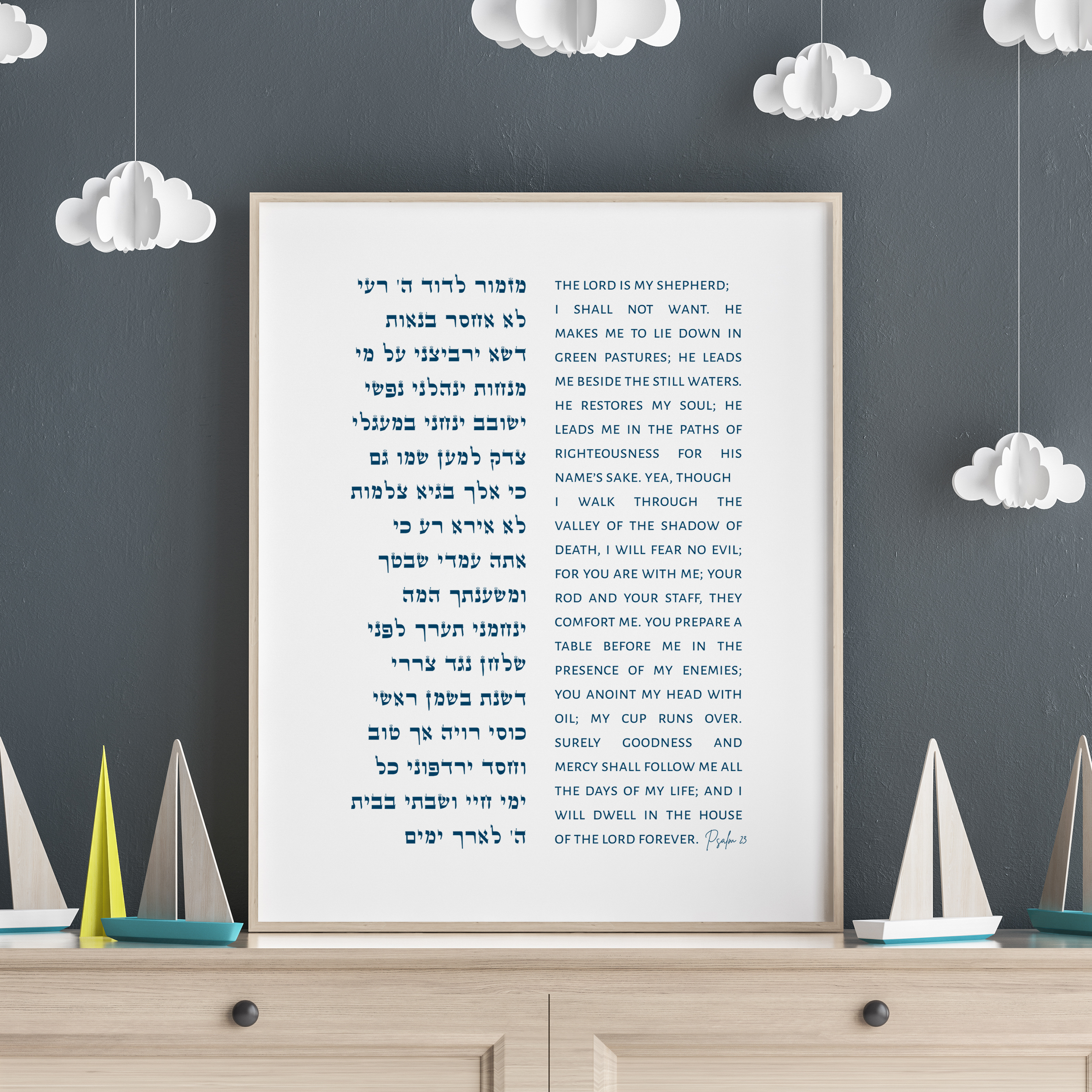 A framed Hebrew and English Bible print of psalm 23 displayed on a light wood dresser against a dark gray wall decorated with white paper clouds.